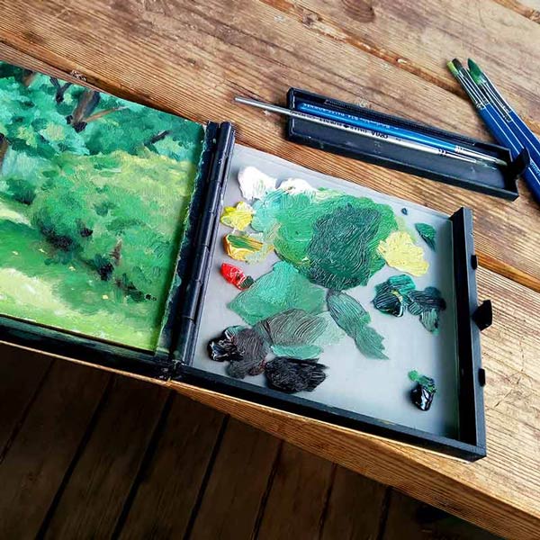 The pochade box is in use and lying flat on a table and you can see a plein air artwork in progress, as well as the mixed oil colours on the palette and also some brushes in a black tray on the table